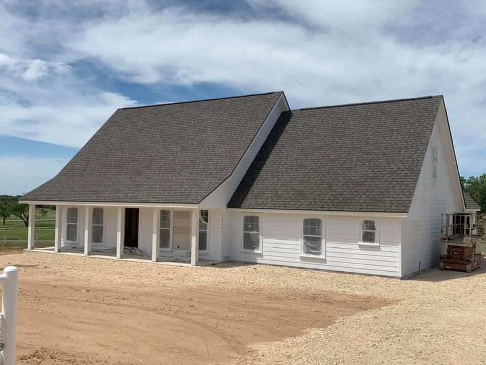 White house with gray roof, covered porch, windows, on gravel. Blue sky background.