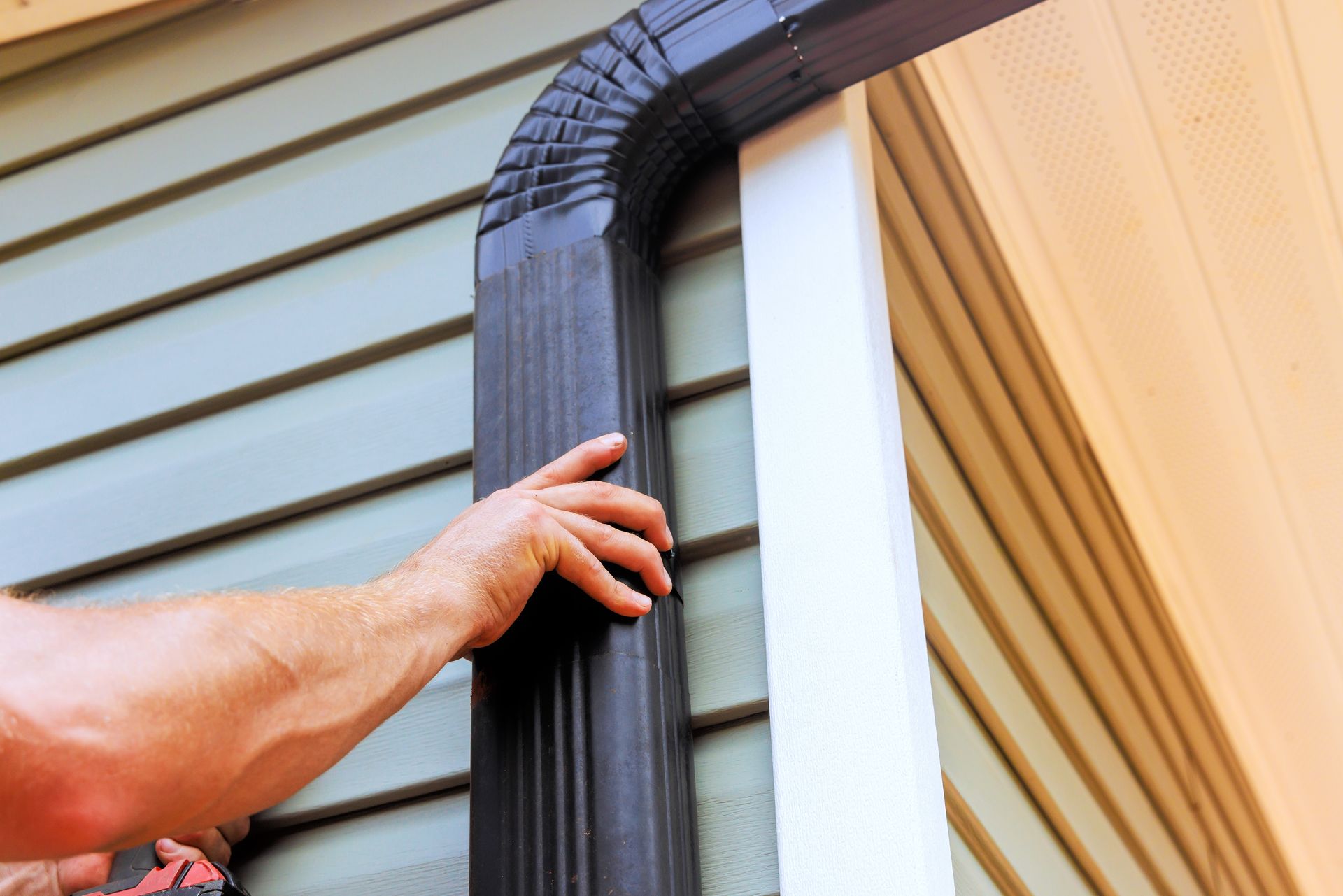 Person installs black gutter on house with green siding.