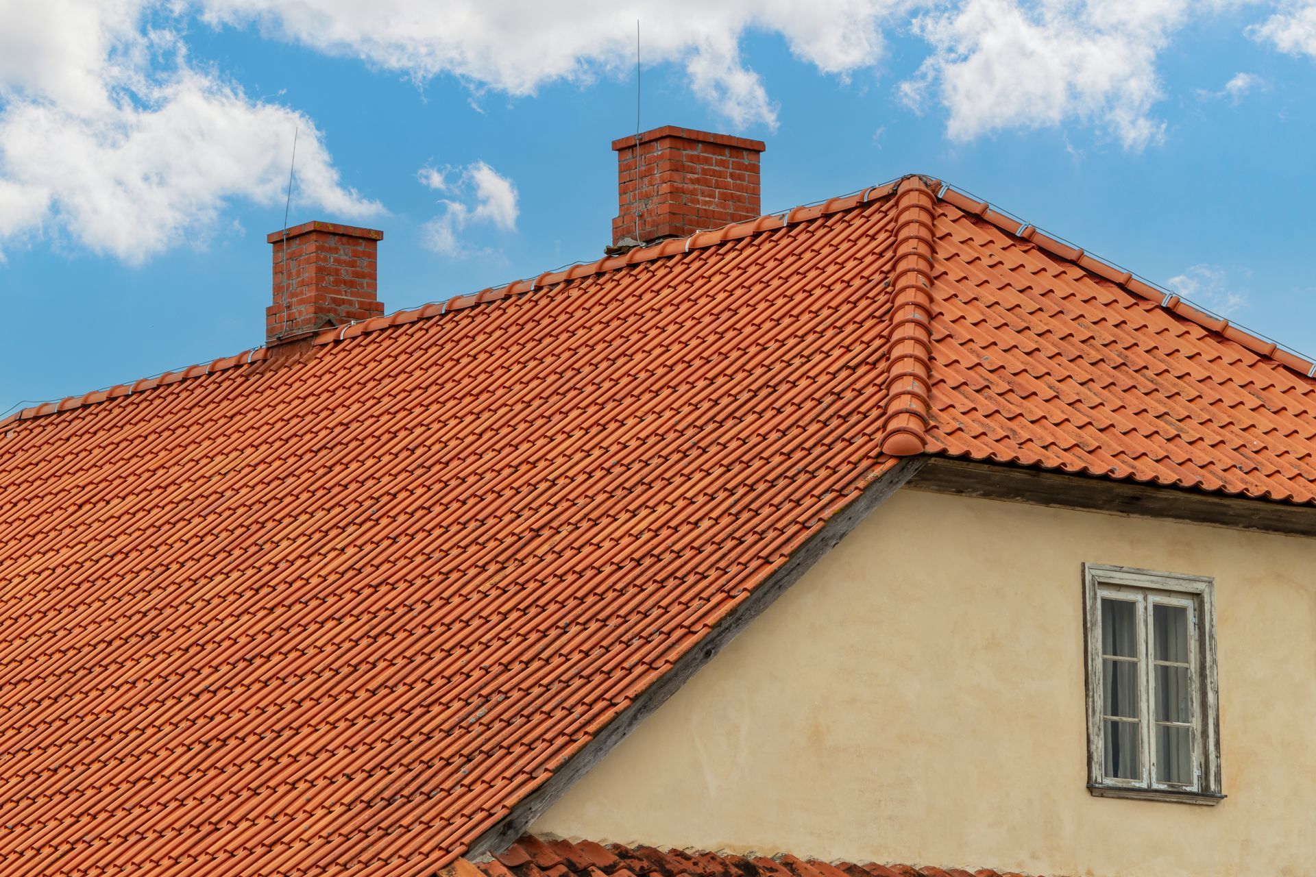 Red tiled roof with brick chimneys against a blue sky, beige building with window.