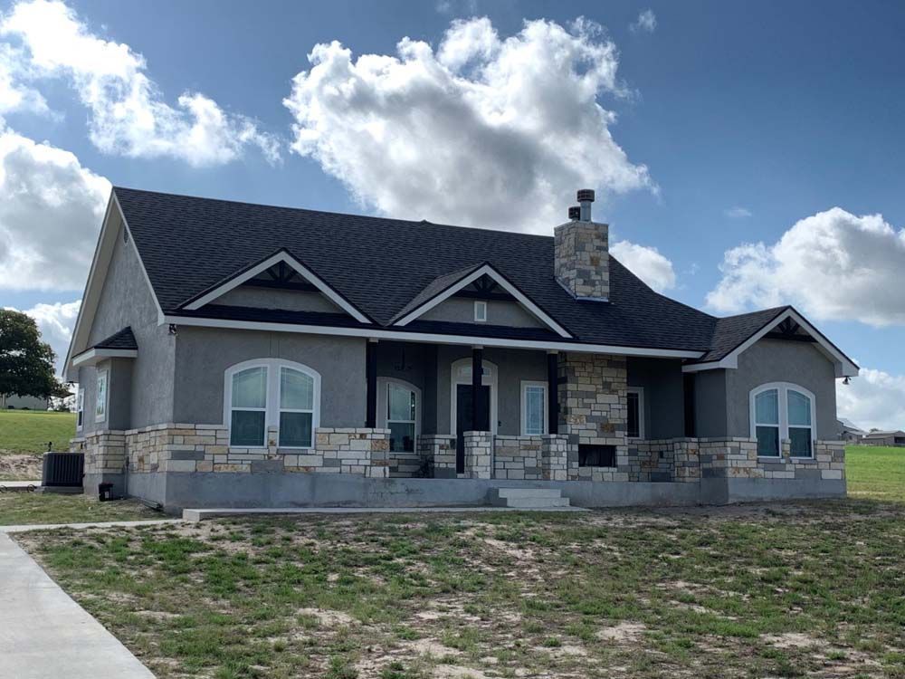 Gray stucco house with stone accents, black roof, and chimney against a blue sky with clouds.