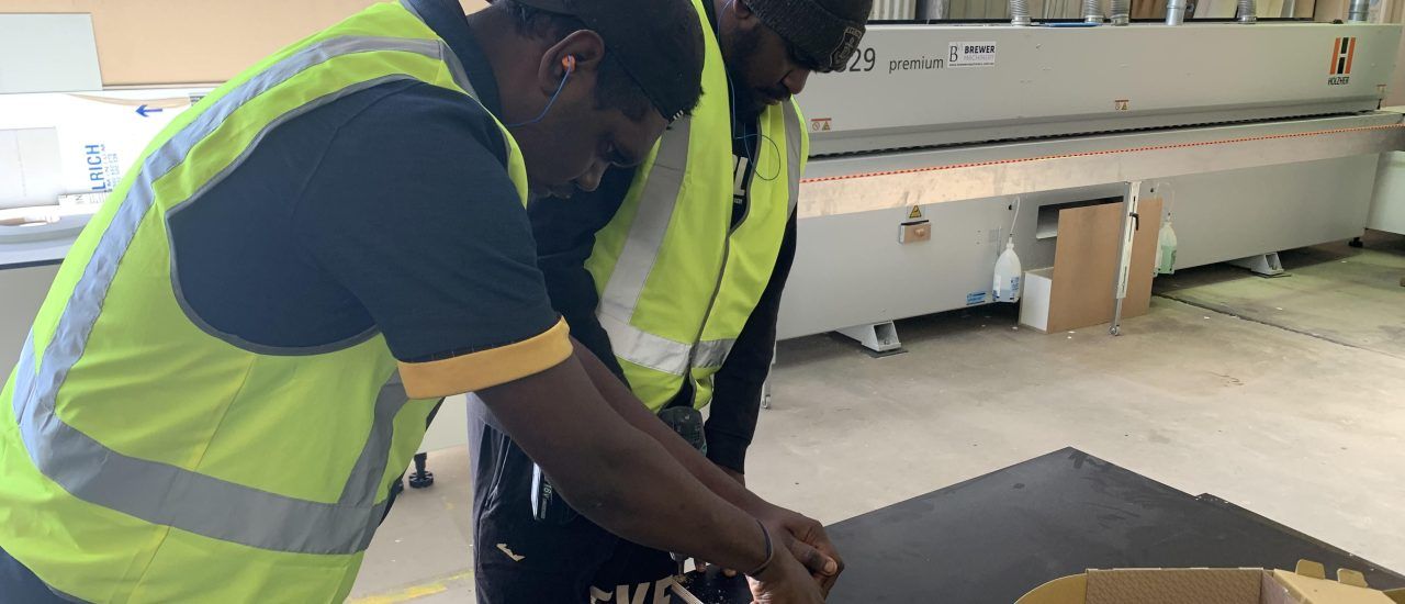 Two men in yellow vests are working on a piece of wood.