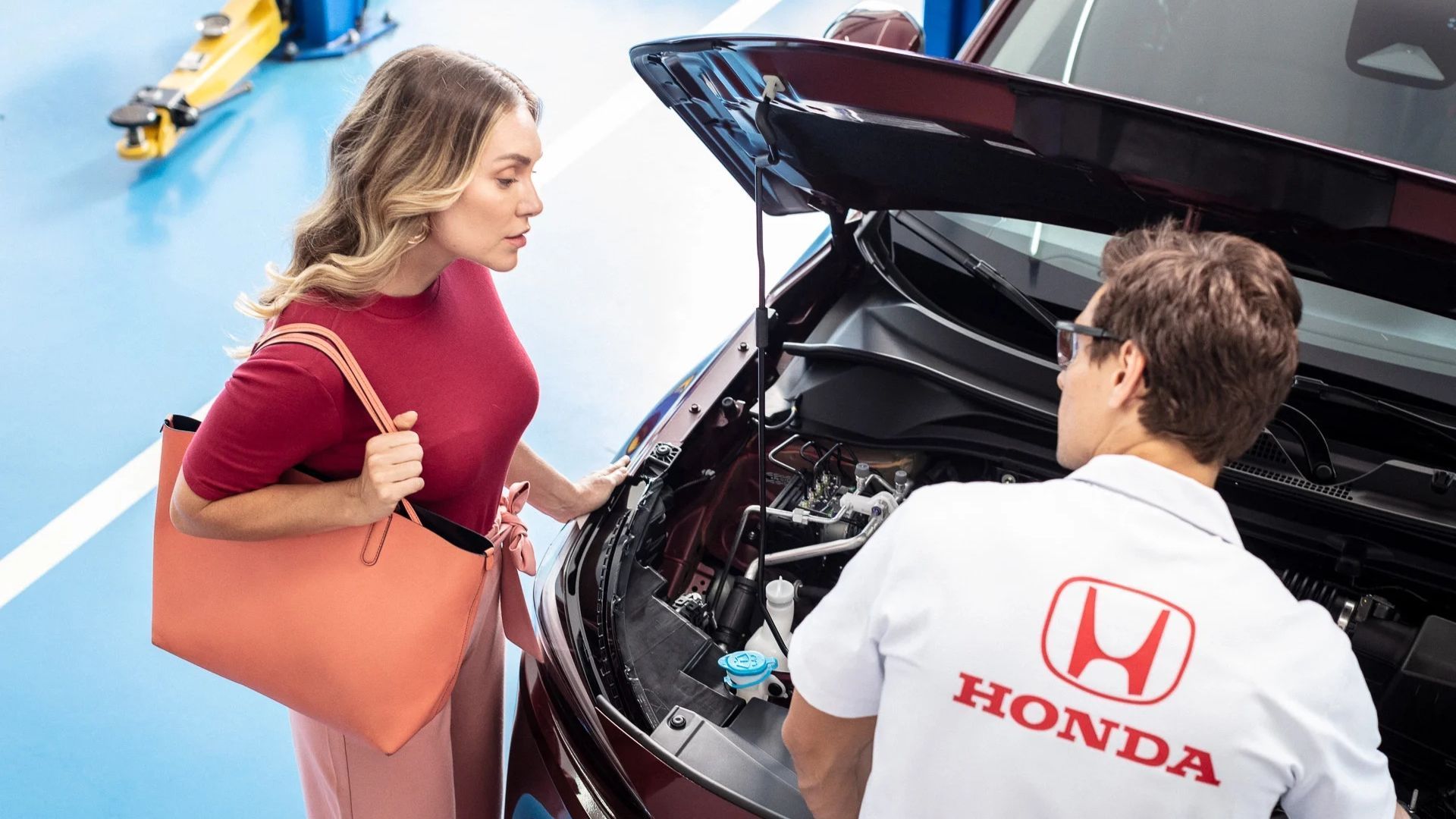 Woman speaking to a Honda mechanic beside an open car hood on a blue roadside.