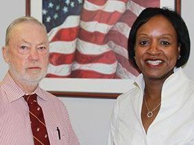 Two people standing in front of a framed American flag, both smiling at the camera.