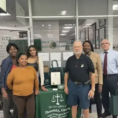 Six people posing outside a storefront, with a green table and sign reading