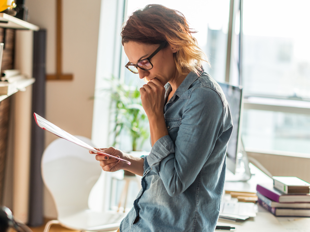 Woman in glasses reading a document in a bright office, standing beside stacked books and a desk