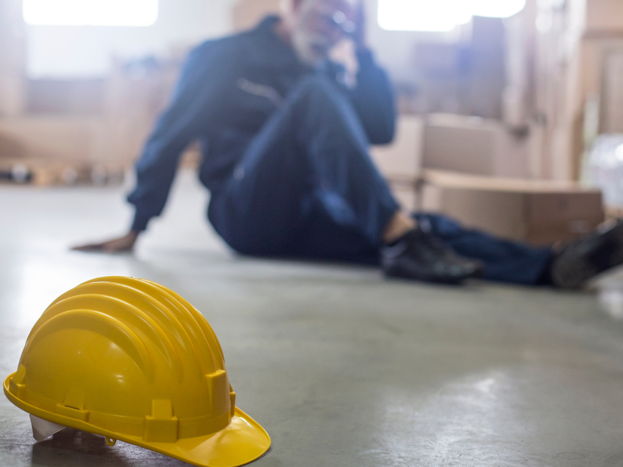 Yellow hard hat in foreground with an injured worker blurred on the floor in a warehouse area