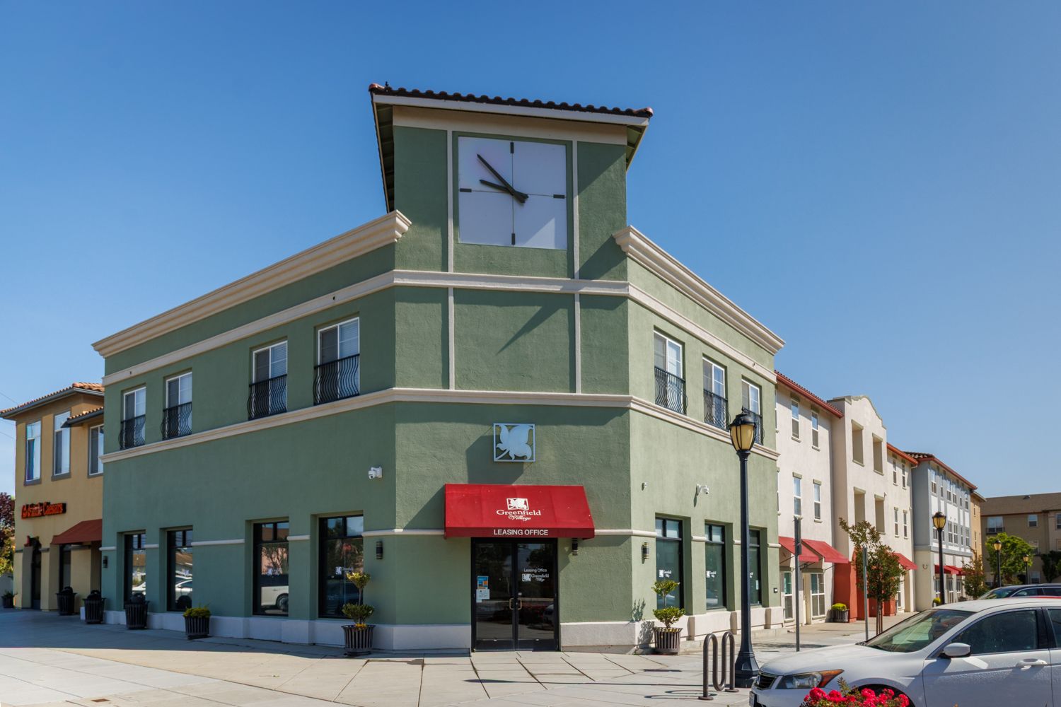 Corner storefront building with clock tower, green facade, and red awning on a sunny street corner