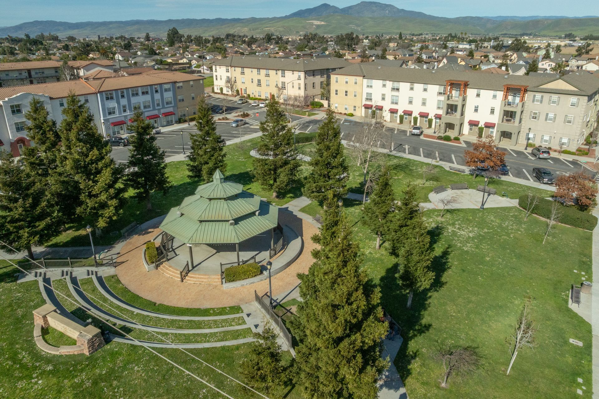 Aerial view of a park with a gazebo, trees, and apartment buildings under a clear sky.