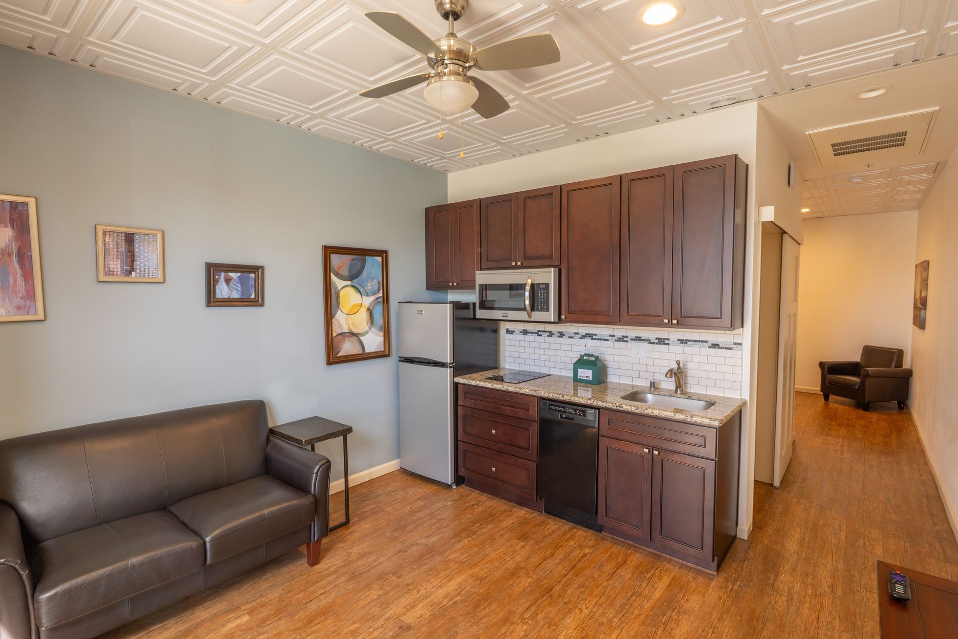 Apartment with dark sofa, kitchenette, wood floors, and a hallway leading to another room
