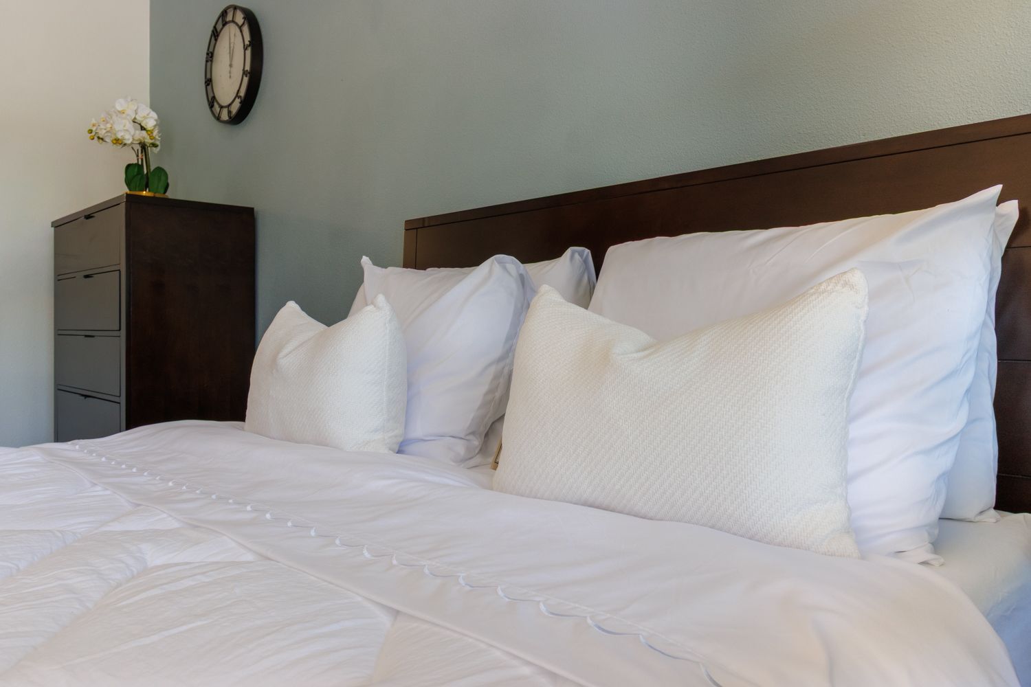 White bed with multiple pillows in a softly lit bedroom with a dark wood dresser and wall clock