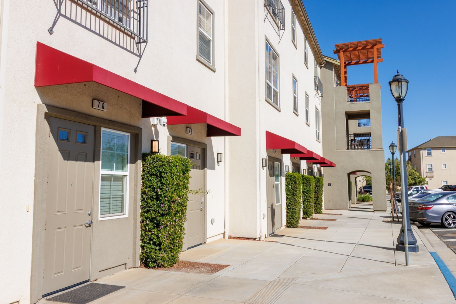 Apartment building sidewalk with red awnings, potted shrubs, and parked cars on a sunny day