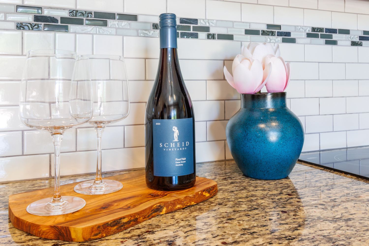 Wine bottle, two glasses, and blue vase with flowers on a granite countertop in a kitchen