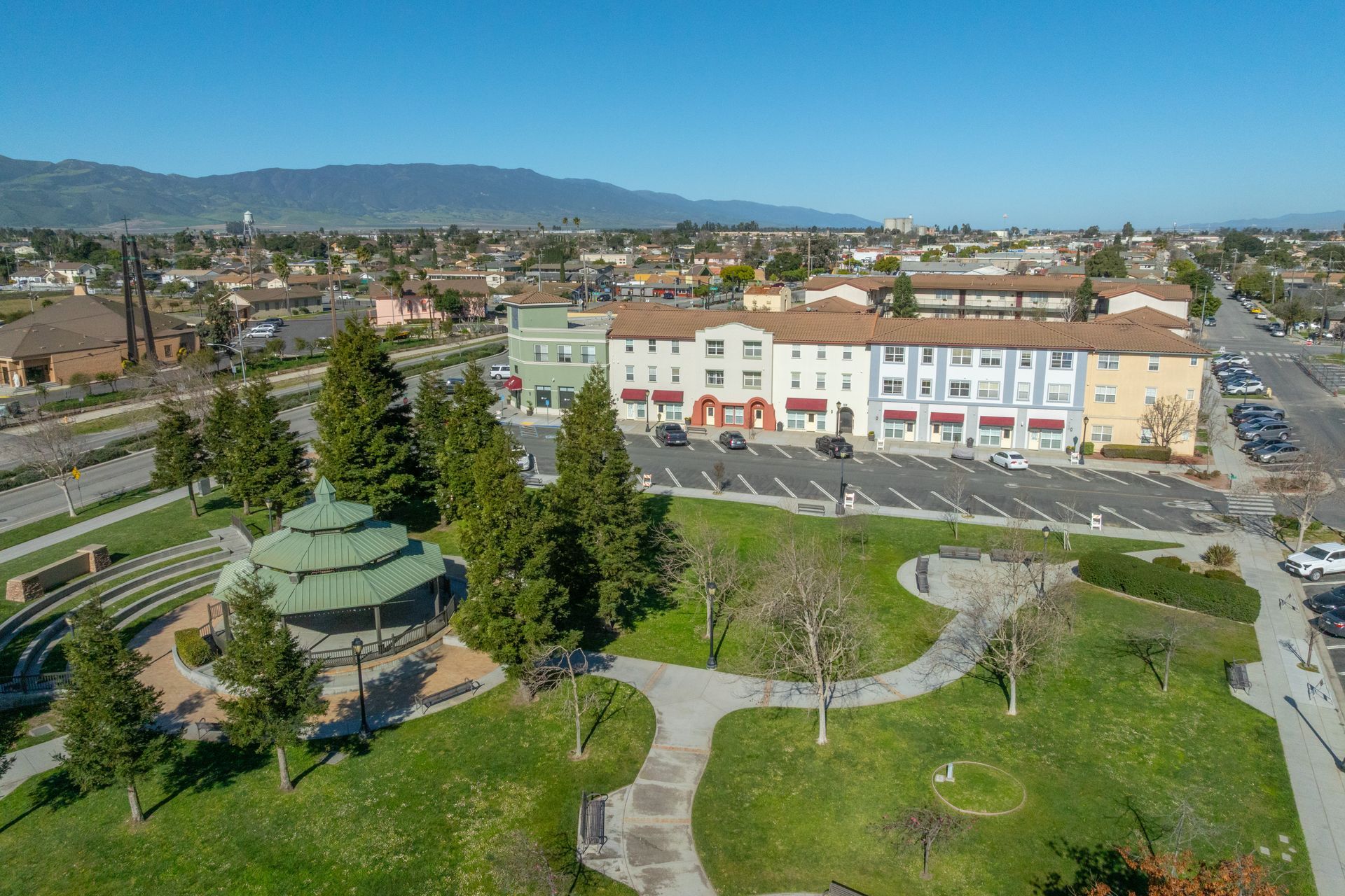 Aerial view of a park with a gazebo, winding paths, trees, and a building under clear skies.