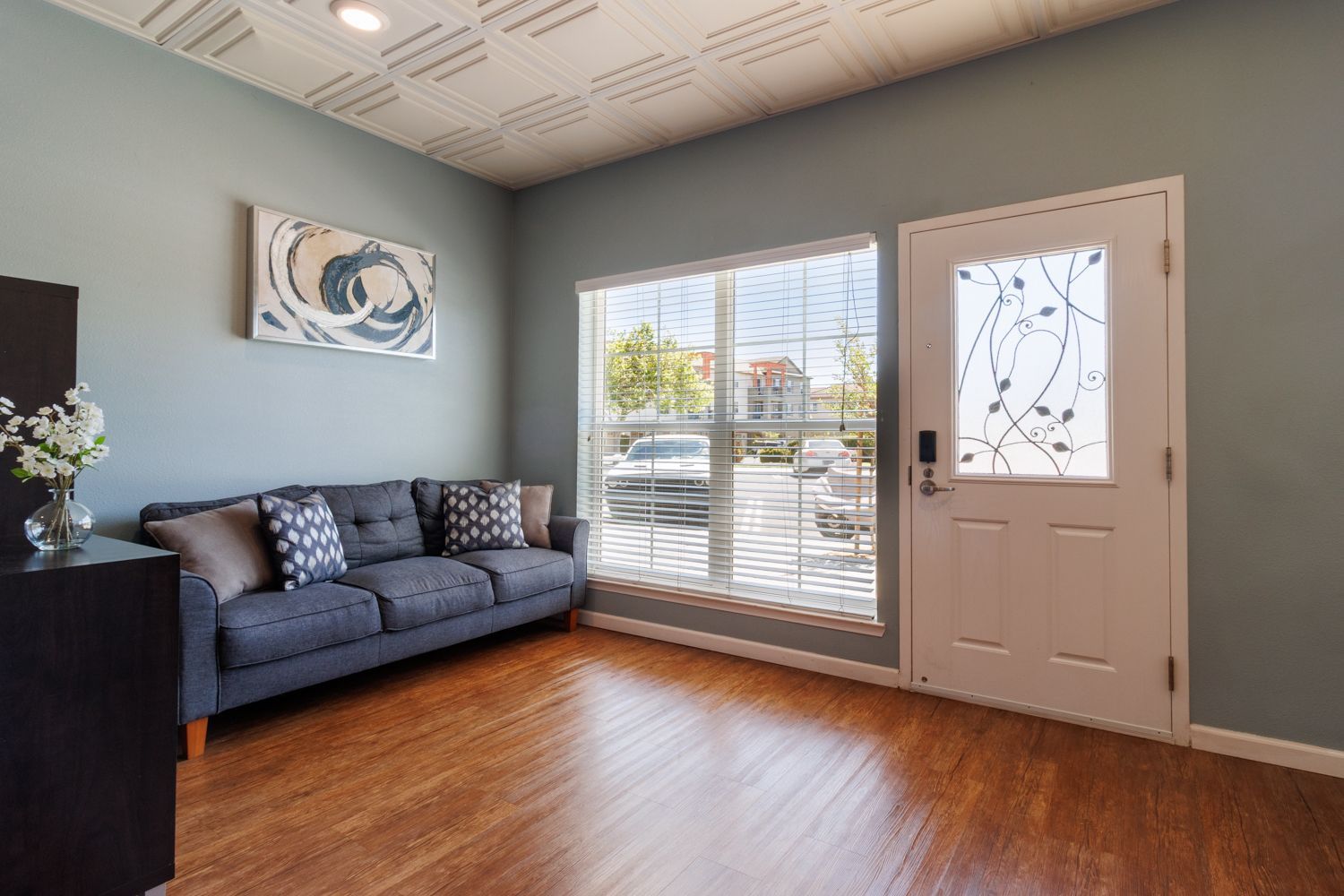 Bright living room with gray sofa, hardwood floor, large window, and white front door.
