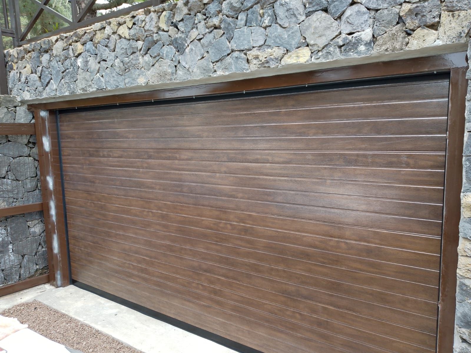 Puerta de garaje de lamas horizontales de color marrón, marco de madera oscura, situada frente a una pared de piedra.