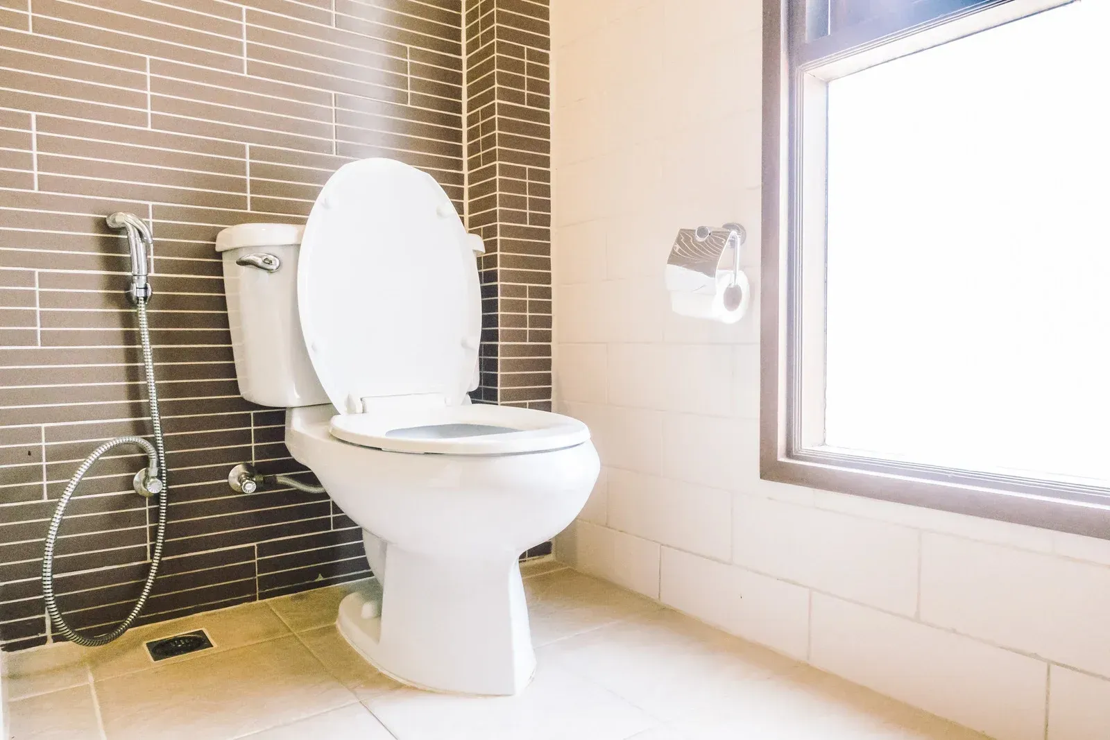 White toilet in a bathroom with brown and white tiles and a window to the right.
