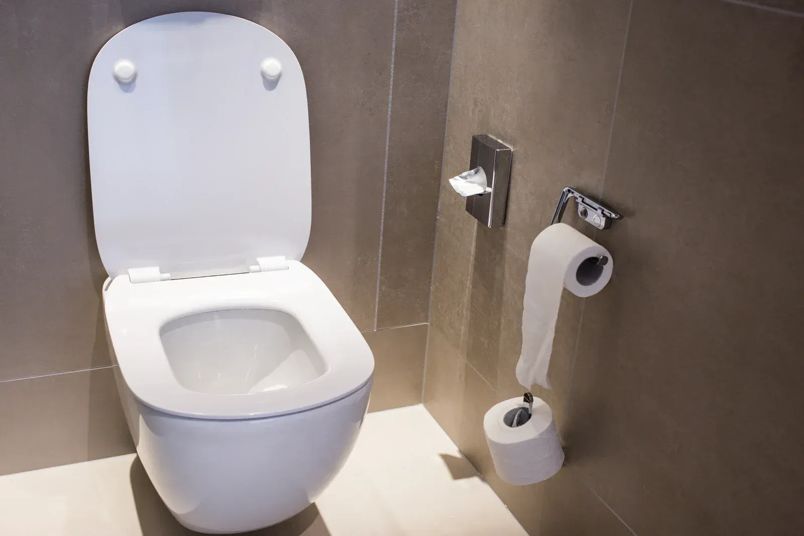 White toilet with the lid up in a bathroom; rolls of toilet paper and shelf above.