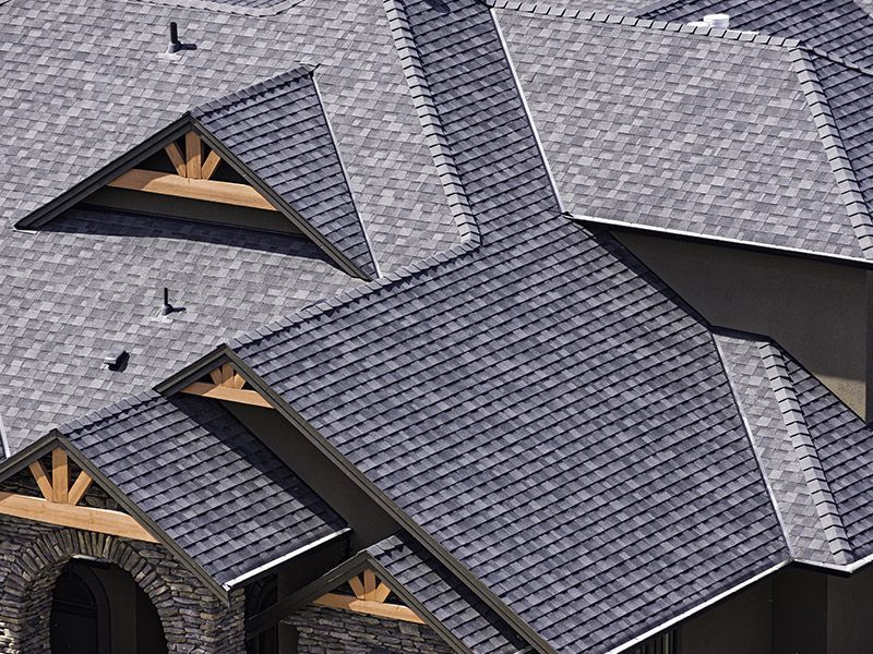 An elevated view of a multi-gabled house roof with dark gray shingles and light brown wood decorative trusses.
