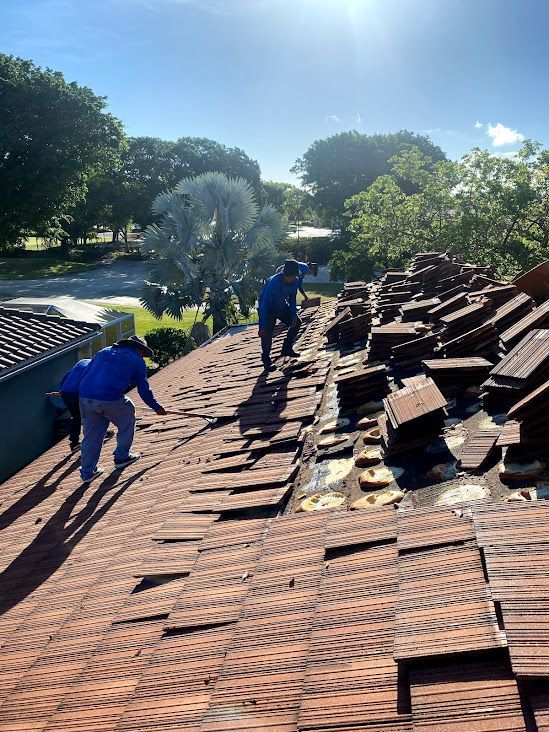 Two workers wearing blue shirts remove reddish-brown roof tiles from a sloped roof on a sunny day.
