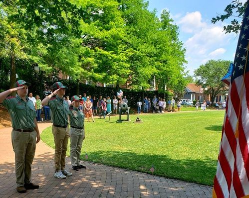 Two uniformed scouts salute on a sunny lawn with a U.S. flag at right.