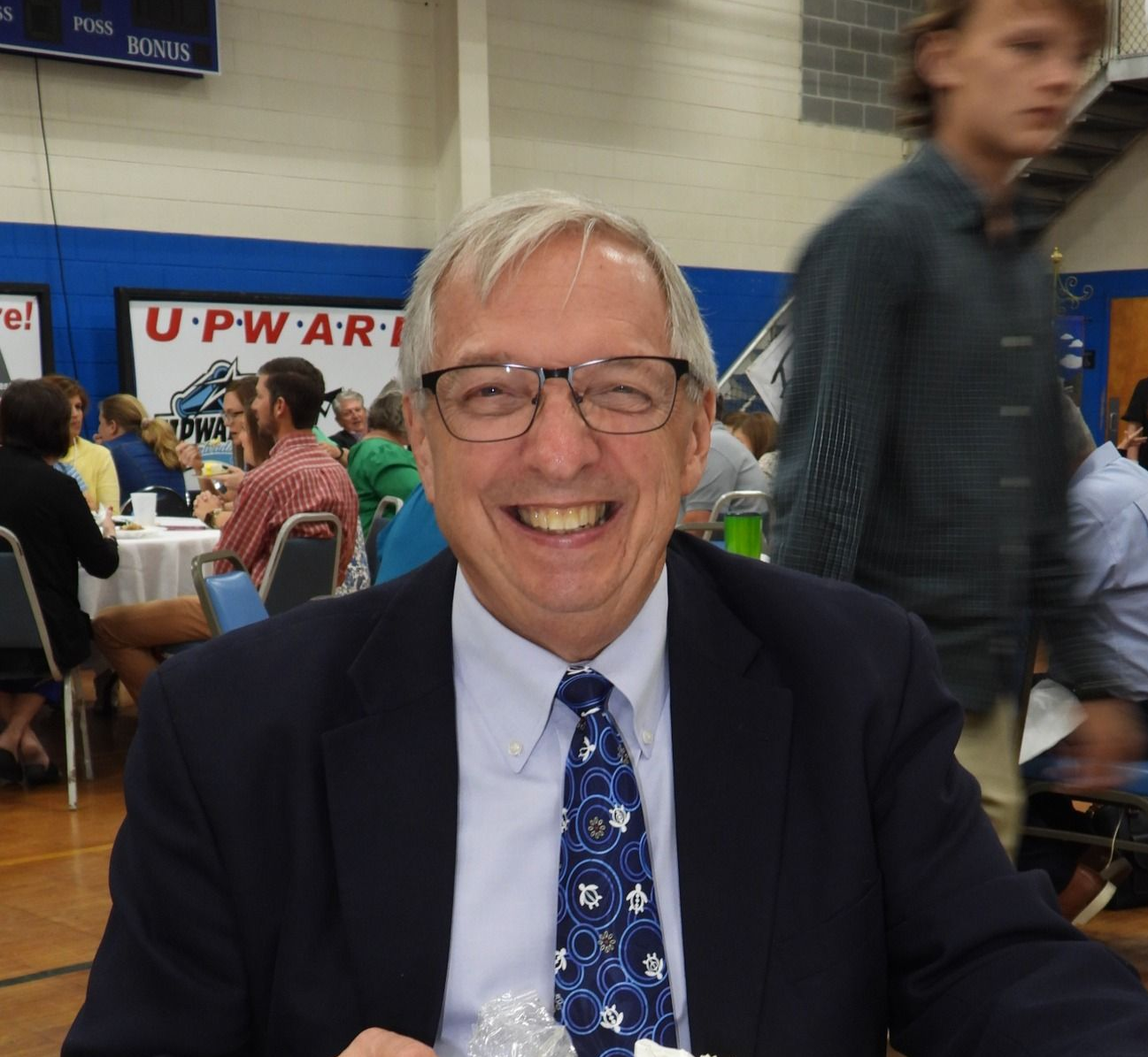 Smiling man in glasses and a blue suit at a crowded indoor event table
