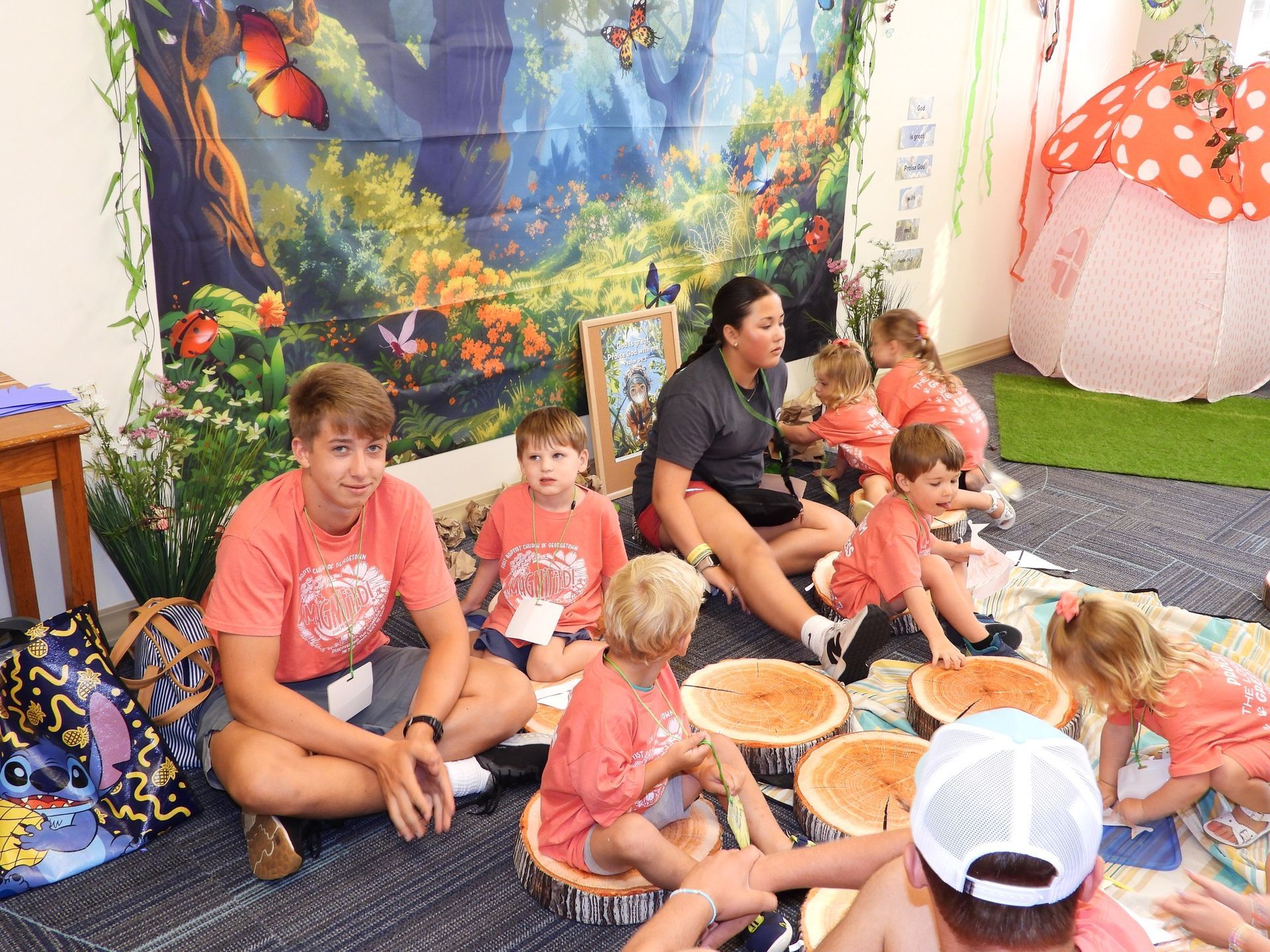 Children seated on a mat at a colorful indoor play area, eating from bowls with an adult nearby
