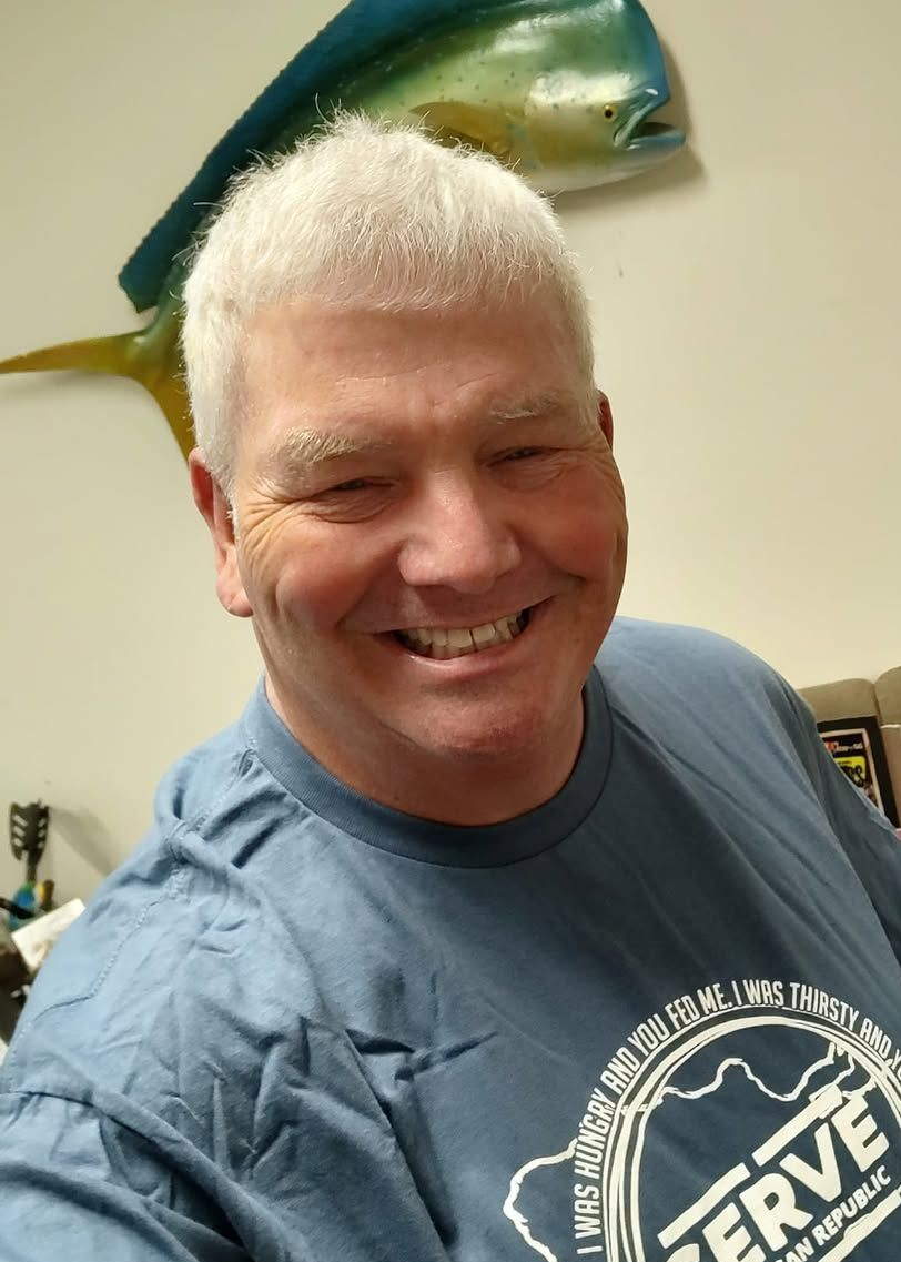 Smiling man in a blue T-shirt indoors, with a large fish sculpture on the wall behind him