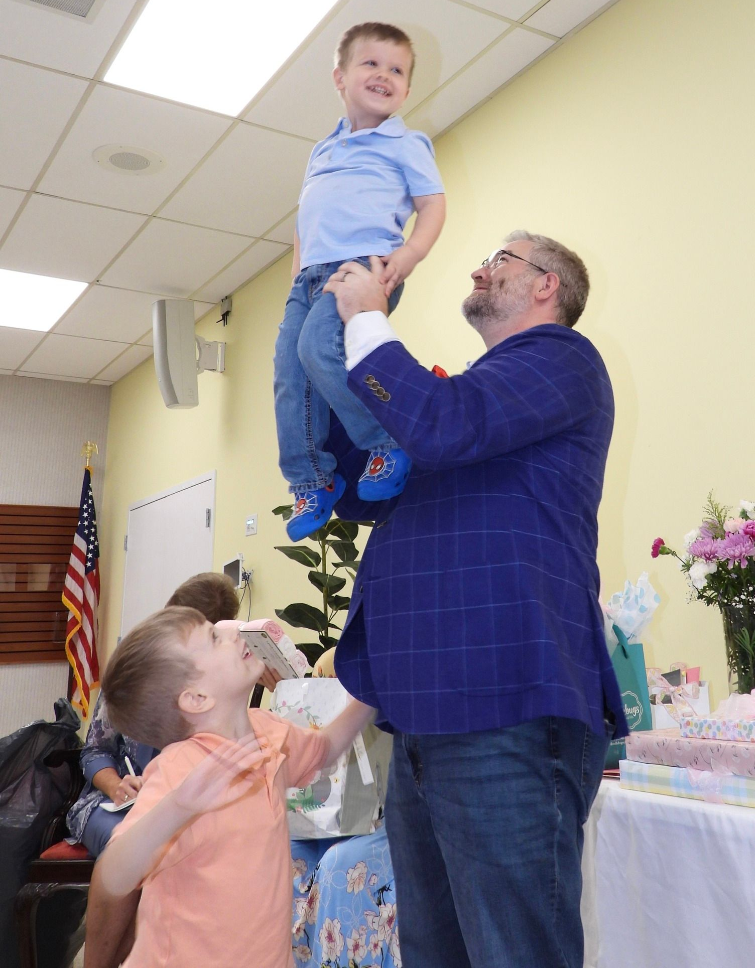 Man holding a smiling child aloft at a decorated indoor event, with children clapping nearby.
