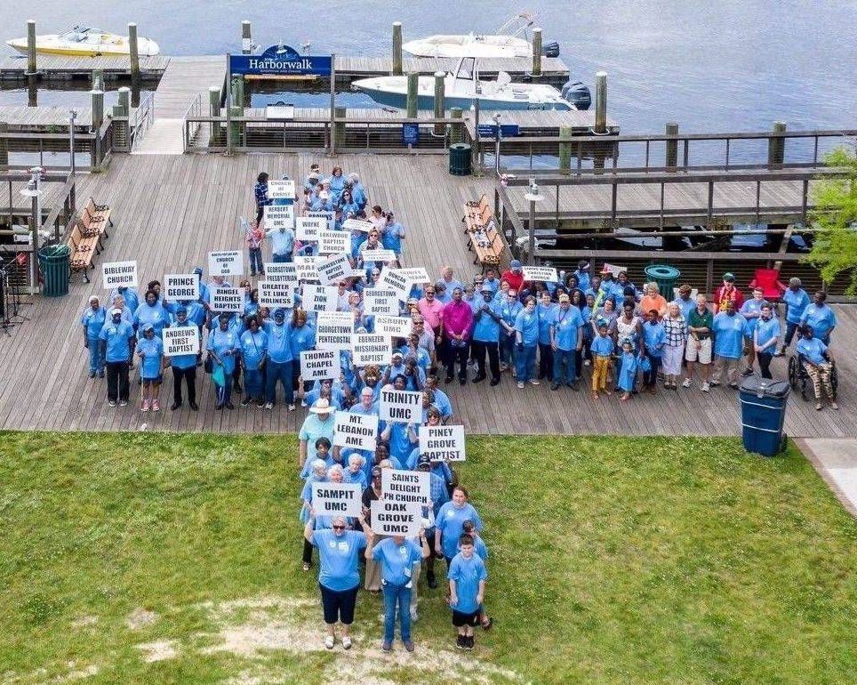 Large group in blue shirts holding signs at a waterfront dock near boats and a grassy shore