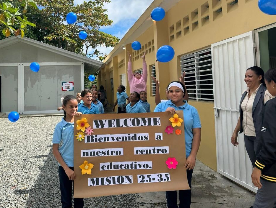 People holding a “Welcome” banner with blue balloons outside a building hallway