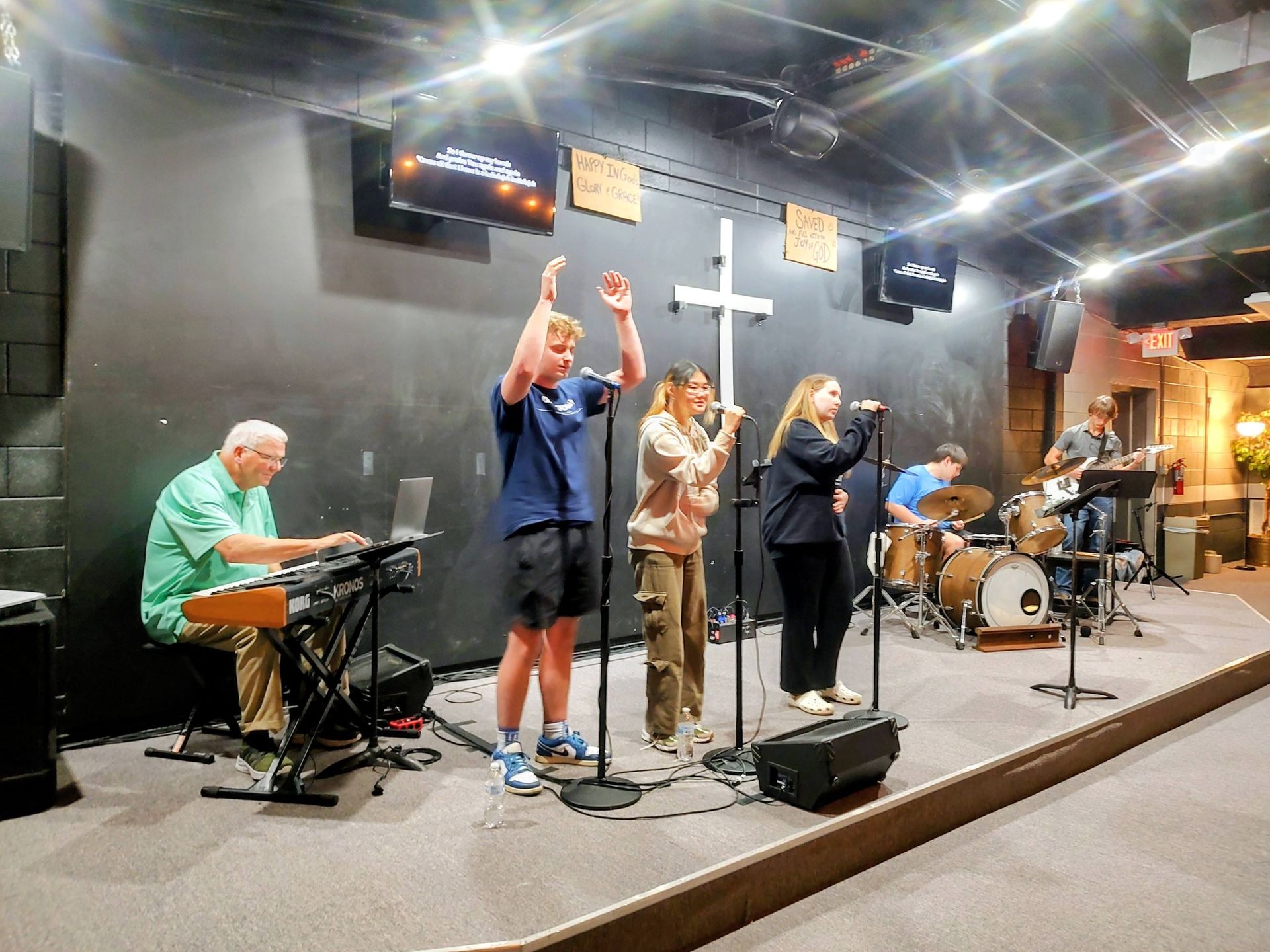 Band performing on an indoor stage, with musicians and microphones under bright lights