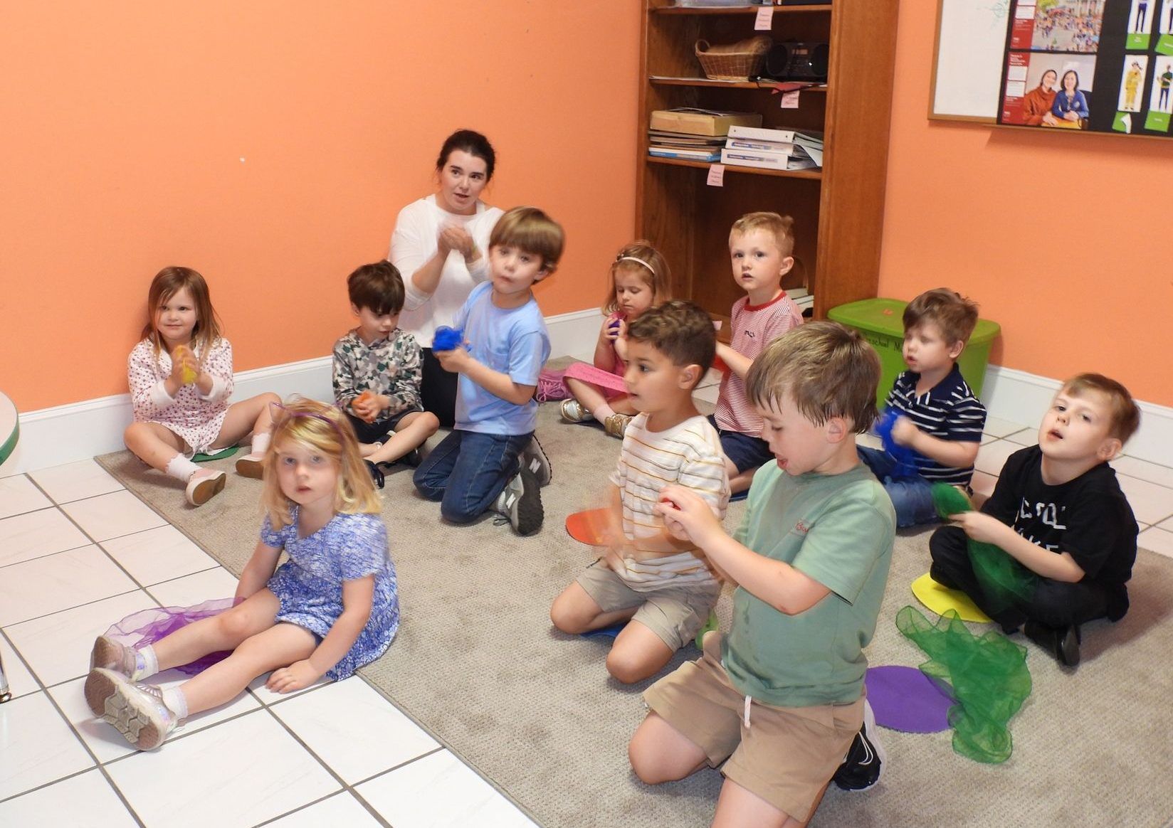 Children sitting on a classroom floor, clapping and watching an activity with a teacher in the background