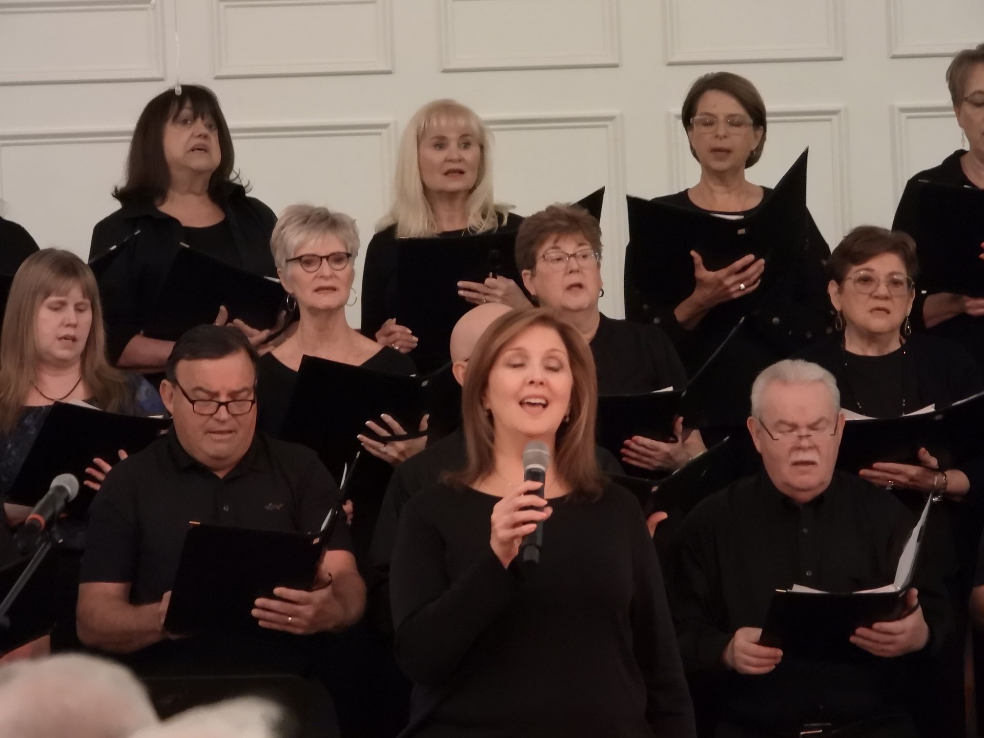 Choir performing indoors, with a woman singing into a microphone in front of seated singers.