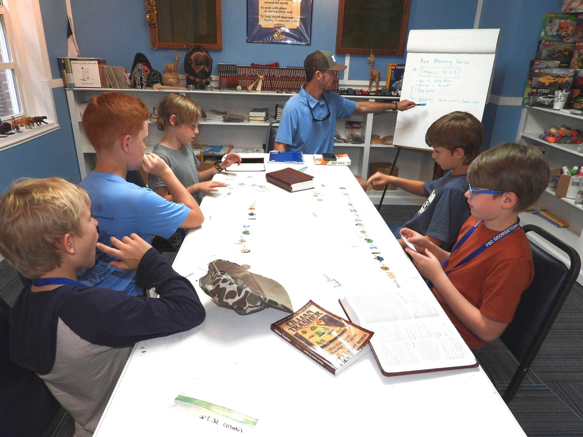 Students seated around a table in a classroom, listening to a teacher at the head of the table.