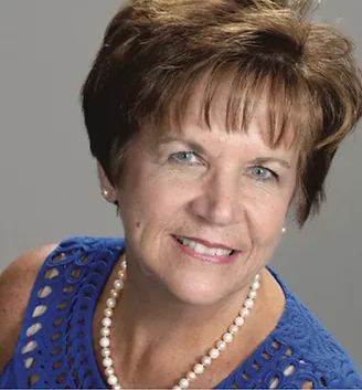 Portrait of a smiling woman in a blue top and pearl necklace against a gray background