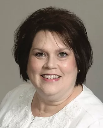 Portrait of a smiling woman with short dark hair wearing a white top against a neutral background