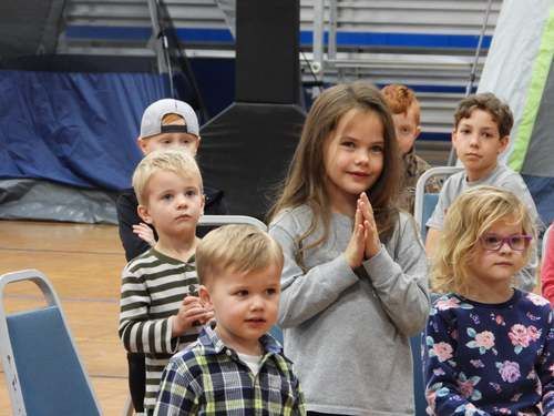 Five children stand indoors on a gym floor, one with hands clasped, all facing the camera.