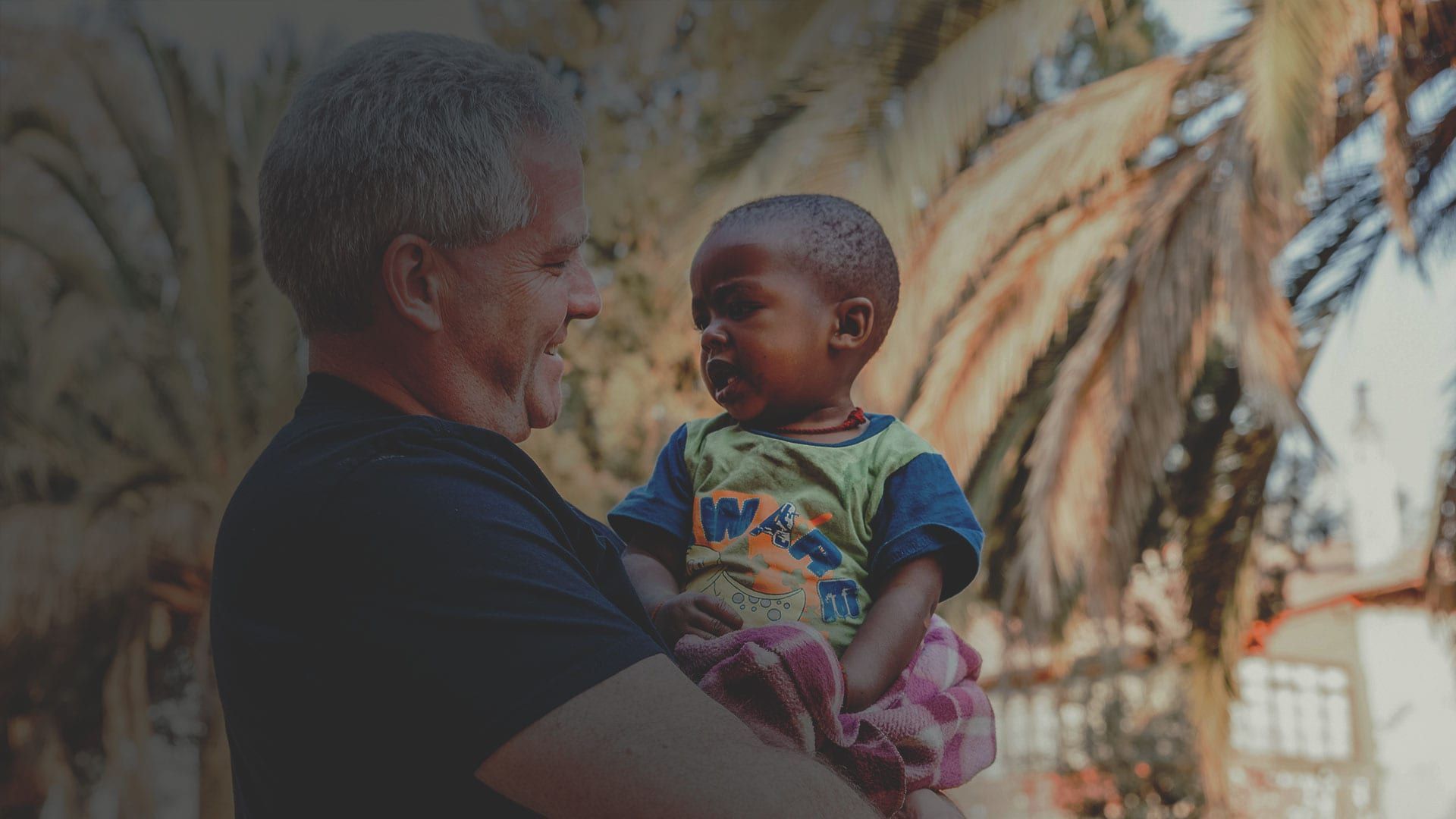Older adult holding a smiling child outdoors near palm trees