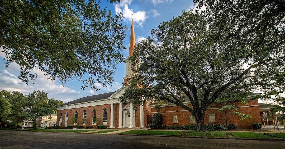 Brick church with a tall steeple under leafy trees on a sunny day.