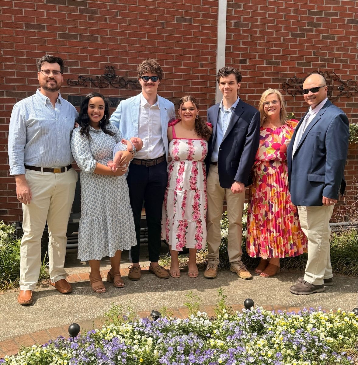 Seven people posing outdoors in front of a brick wall and flower bed, dressed in semi-formal attire.