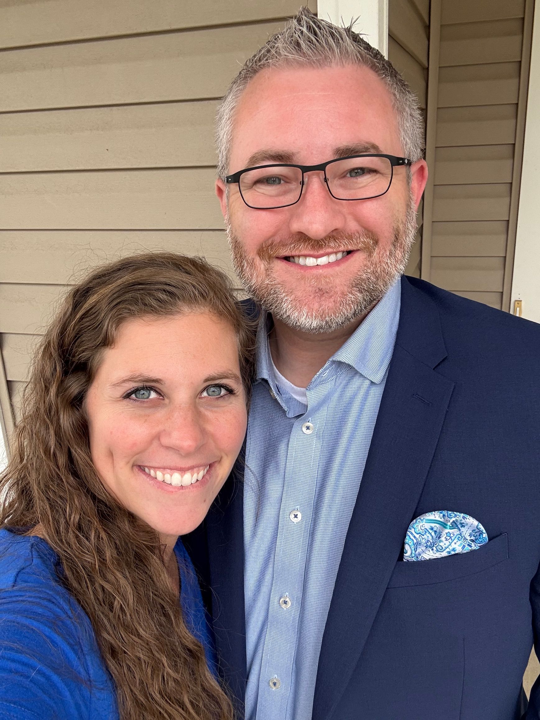 Smiling couple posing on a porch, wearing blue and white, with a tan siding background.