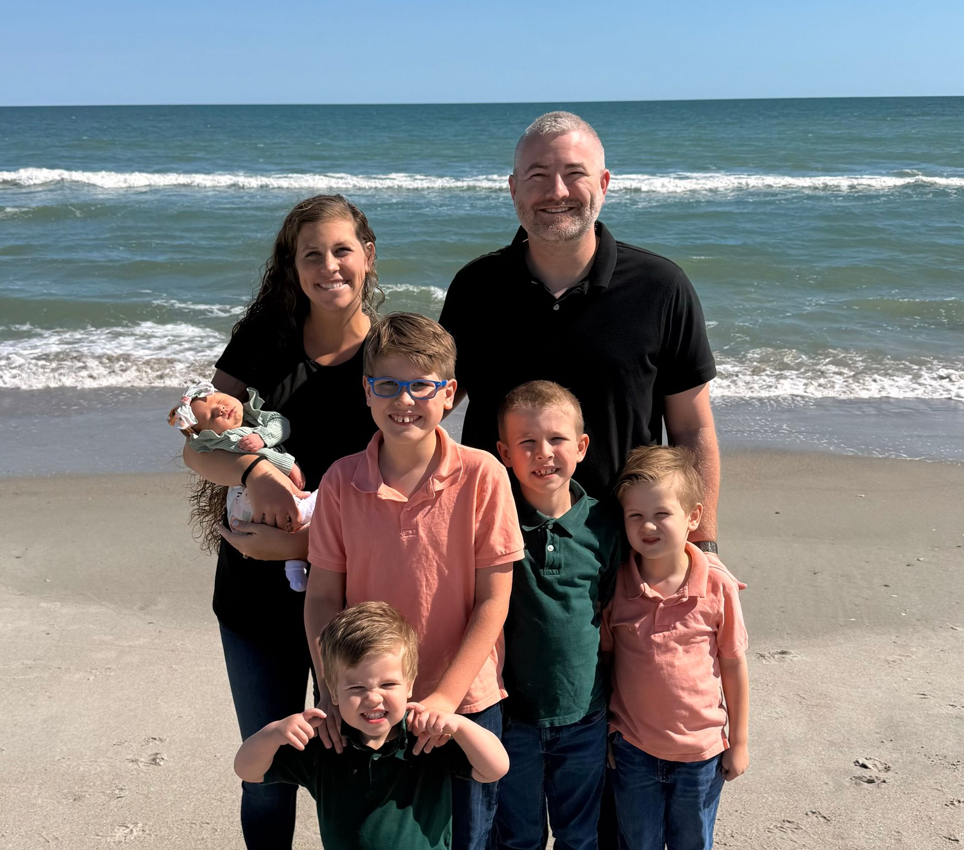 Family of five standing on a sunny beach with ocean waves behind them, smiling at the camera.
