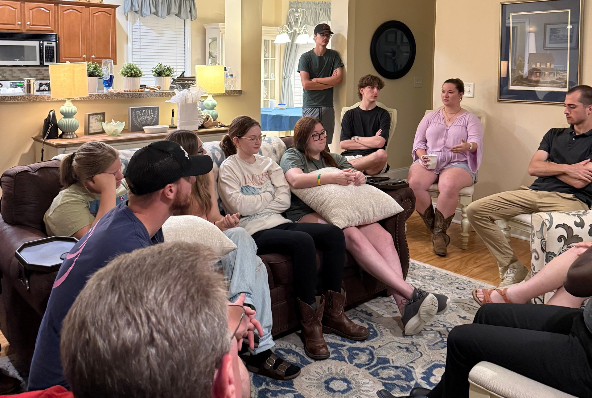 Group seated and standing in a cozy living room during a casual gathering, with kitchen in the background.