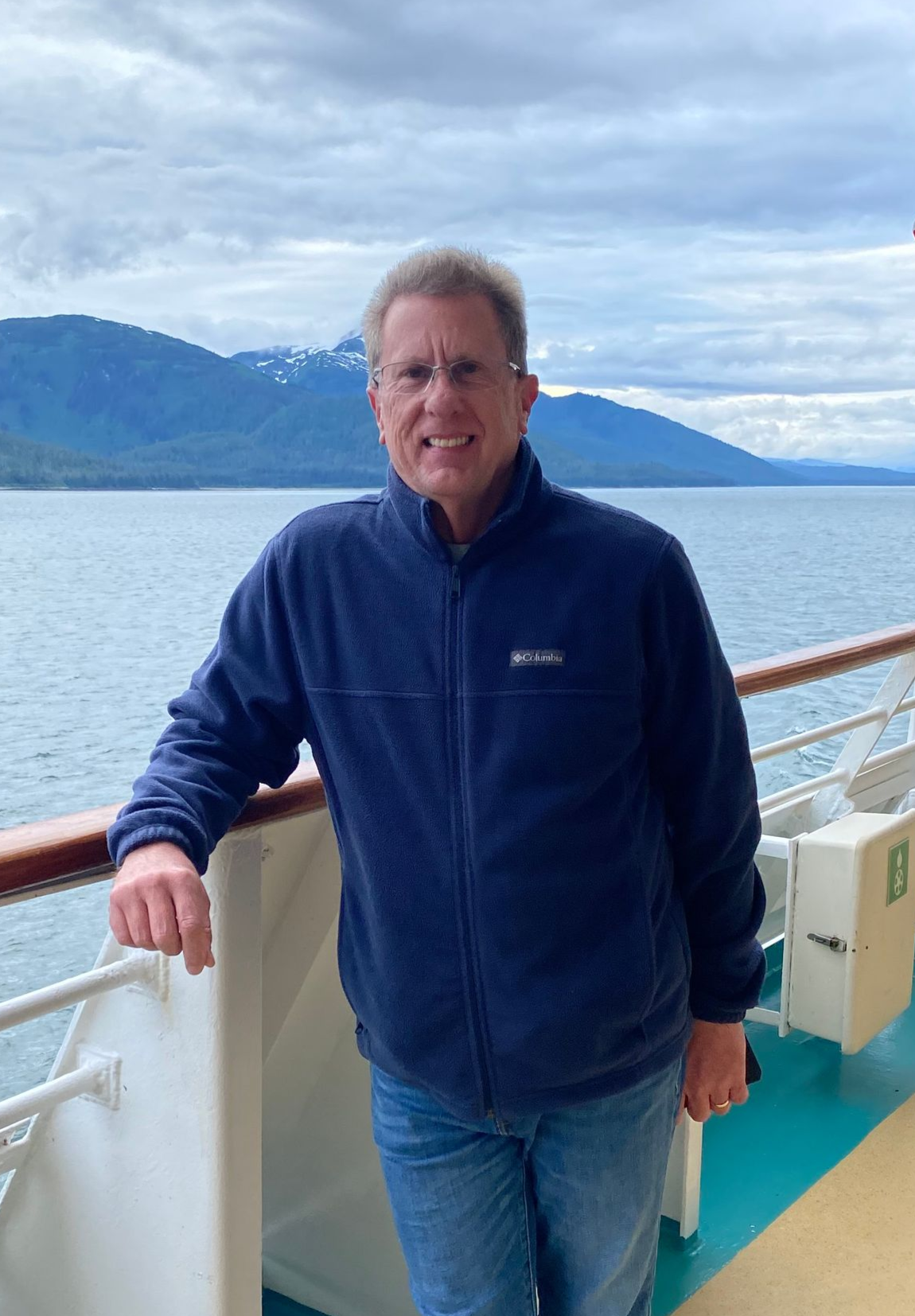 Man standing on a ship deck by the sea, wearing a navy jacket and jeans, with mountains in the background