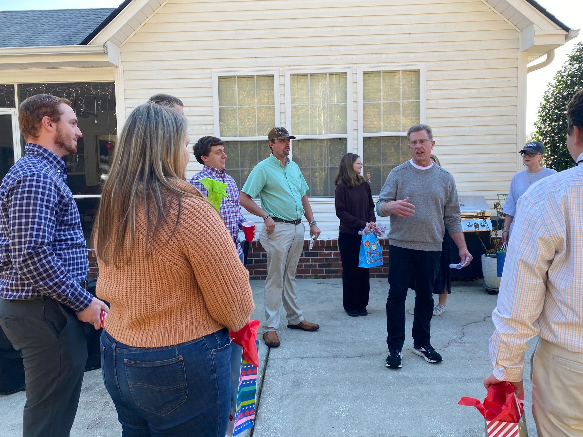 People standing and talking in a driveway outside a beige house, some holding bags and gifts.