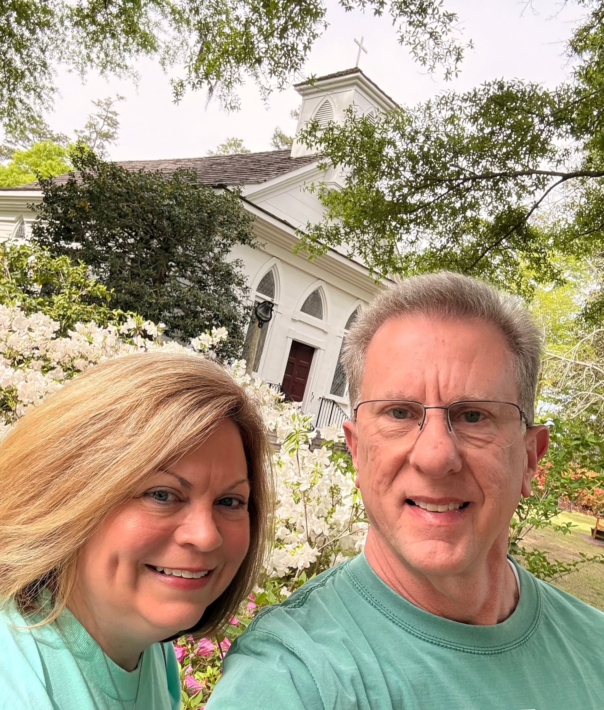 Two smiling people in teal shirts posing outdoors in front of a white building and flowers