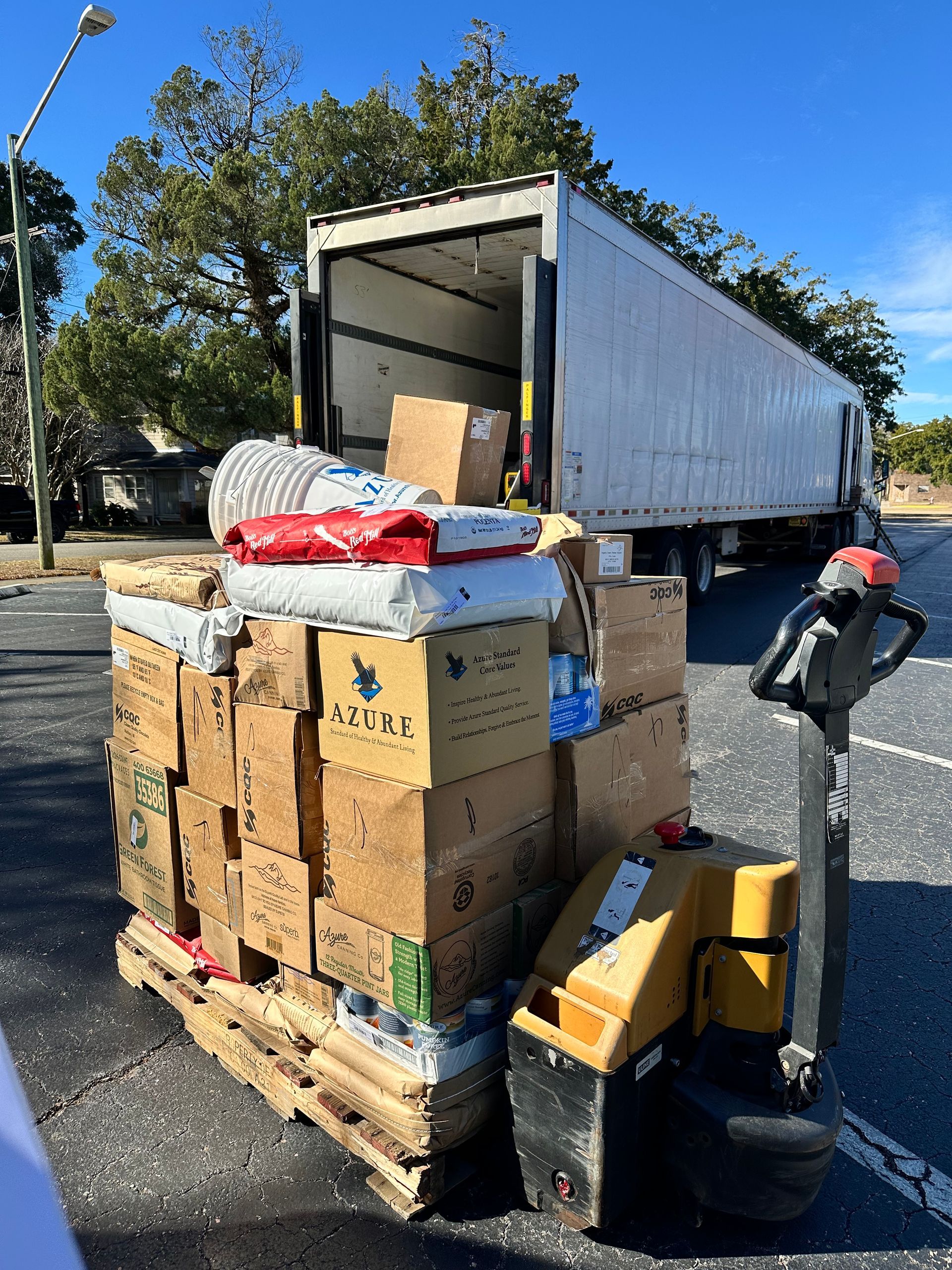 Stacked cardboard boxes and bundled items on a pallet beside an open truck in a parking lot.