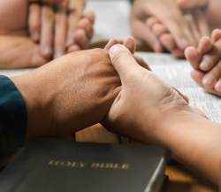 Hands clasped in prayer beside a Bible during a group prayer meeting