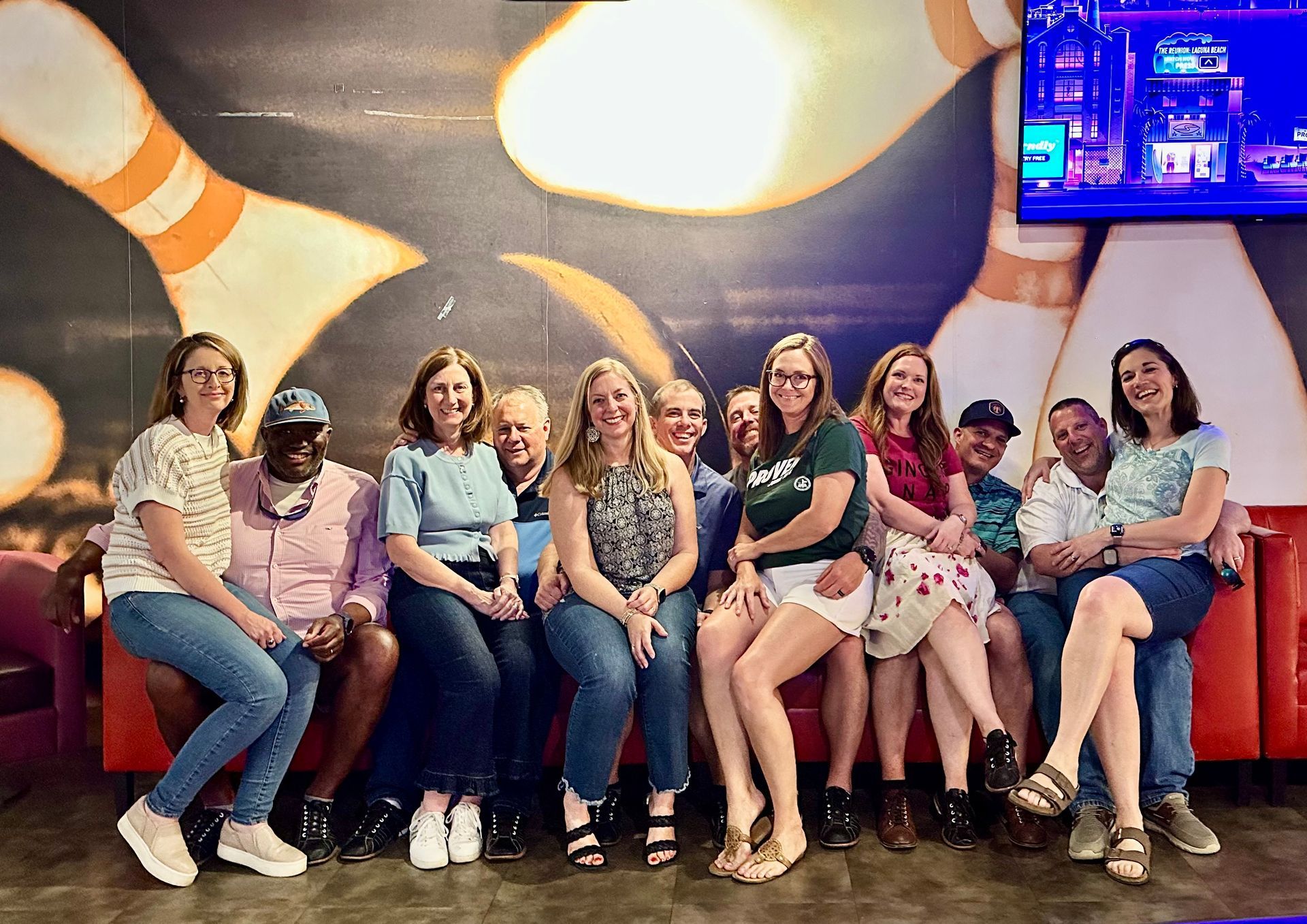 Nine people seated on a red couch in a colorful lounge, posing and smiling under a bright mural.