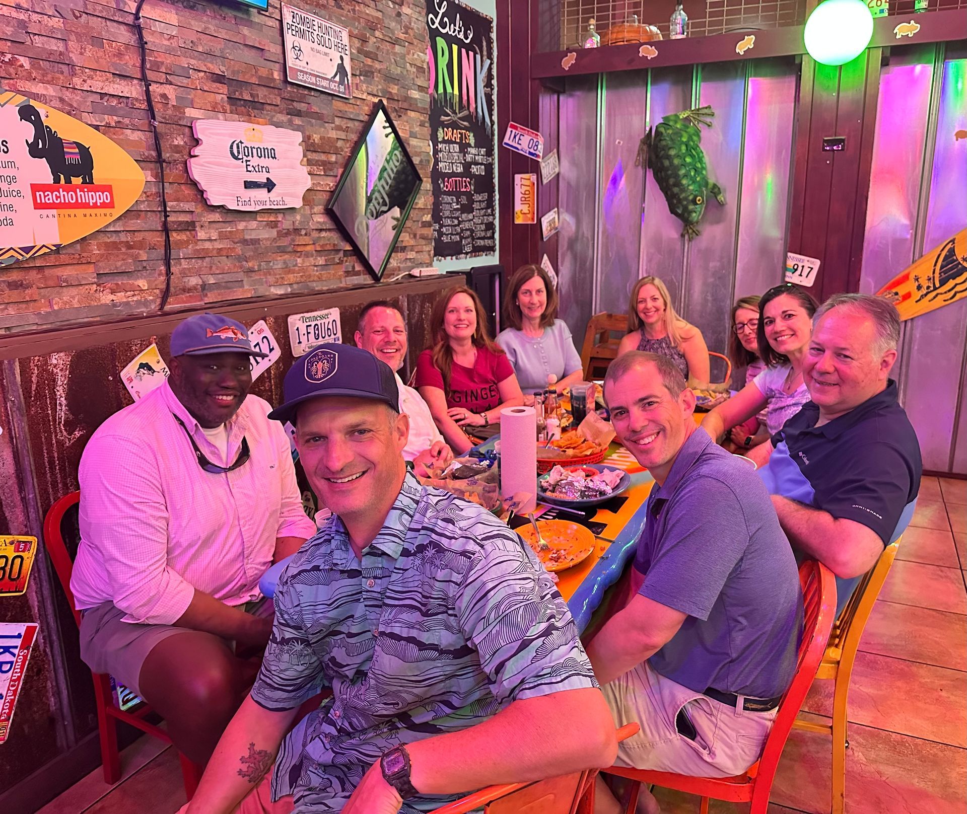 Group of friends seated at a restaurant table, smiling and dining under colorful neon lighting.